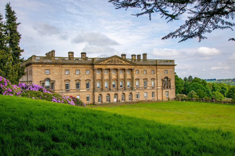 Historic mansion with classical architecture, including columns and many windows, set on a manicured lawn with flowering bushes and trees under a partly cloudy sky.